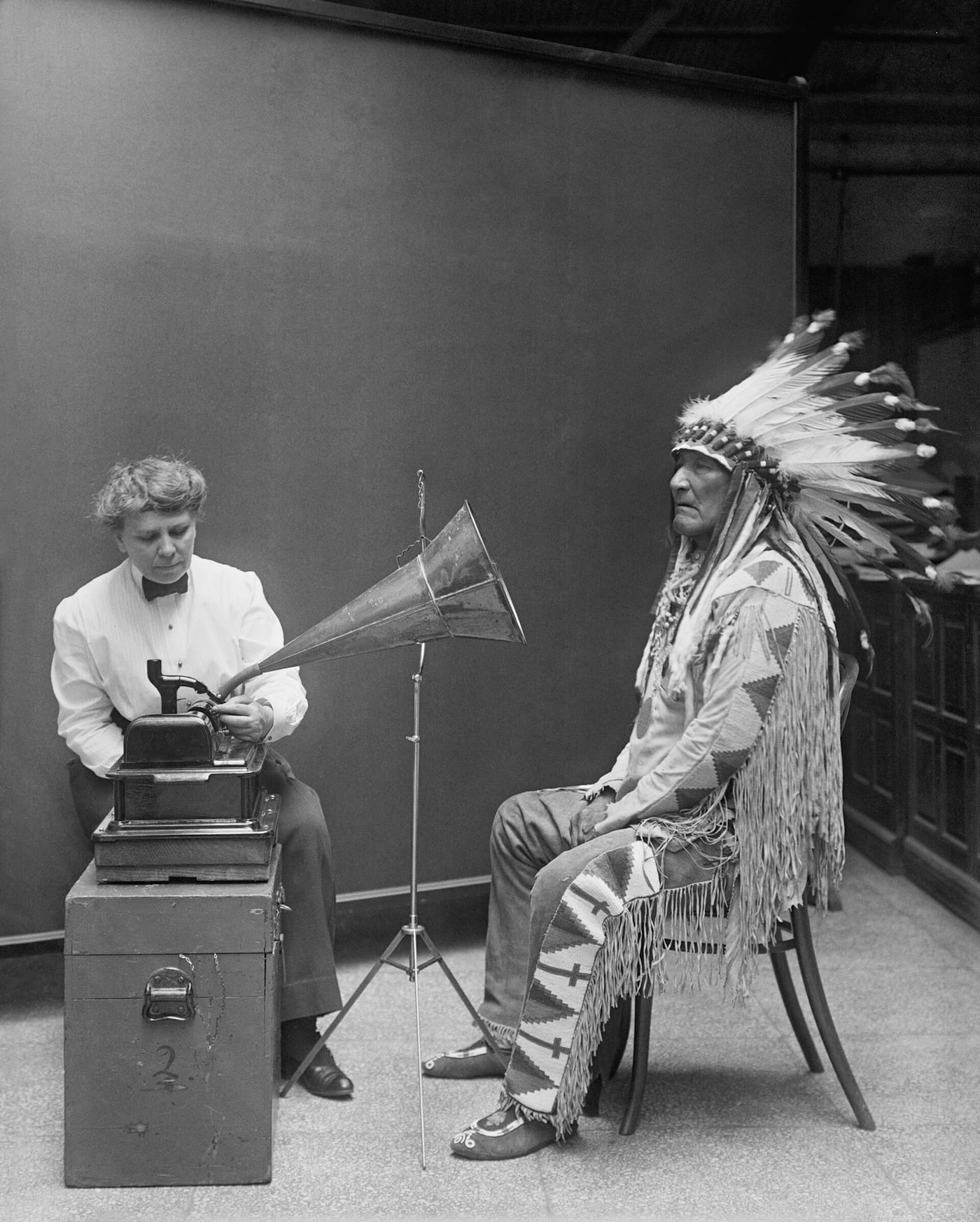 Photograph of Frances Densmore recording Blackfoot chief Mountain Chief on a wax-cylinder phonograph at the Smithsonian in 1916, with a photograph of Mountain Chief in traditional dress behind him.